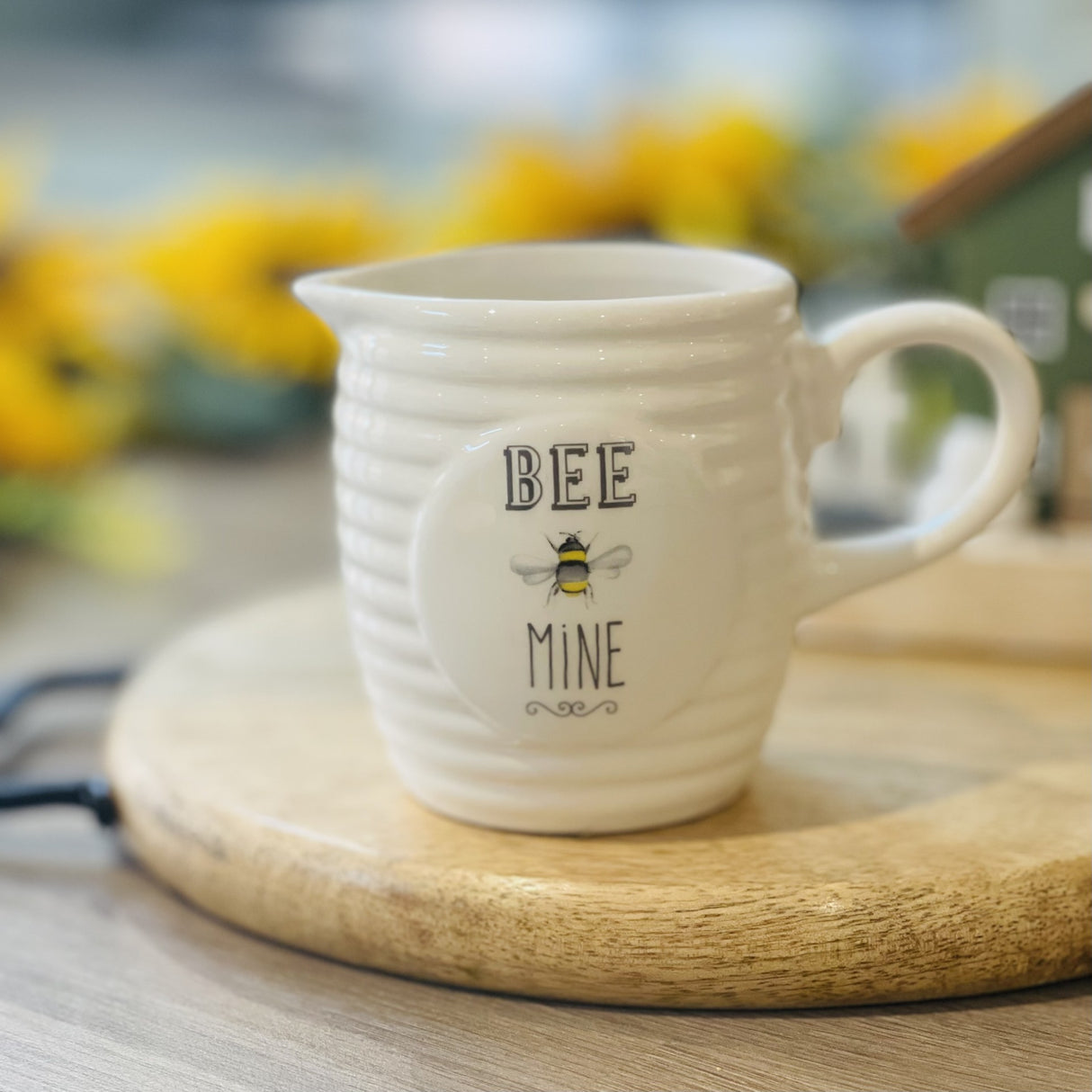 A white ceramic creamer jug with the words Bee Mine on the front, placed on a wooden chopping board