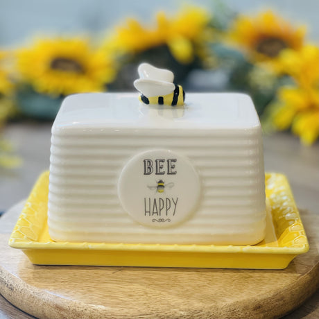A ceramic white butter dish with the words Bee Happy on the front and a ceramic bee on the top