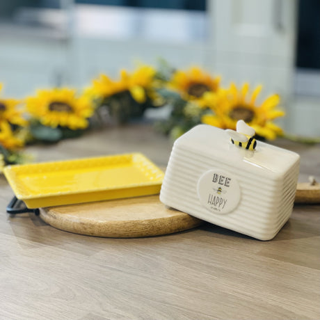 A ceramic white butter dish with the words Bee Happy on the front and a ceramic bee on the top