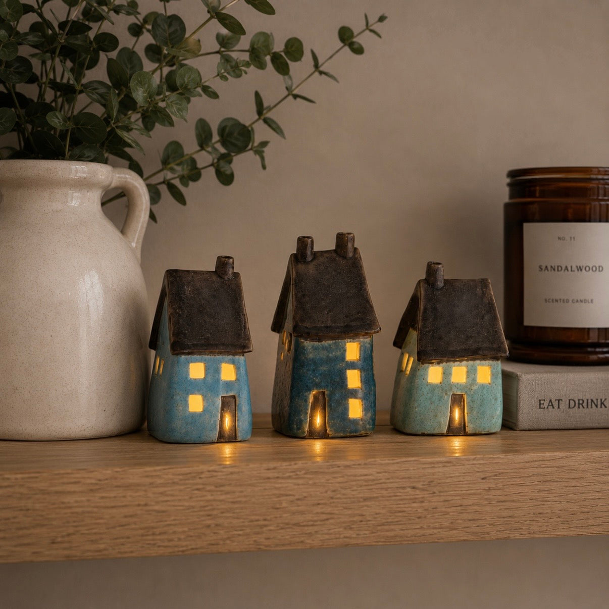 Three small ceramic LED house figurines in blue and turquoise glazes with dark brown roofs, displayed on a wooden shelf next to a white ceramic pitcher and a candle.
