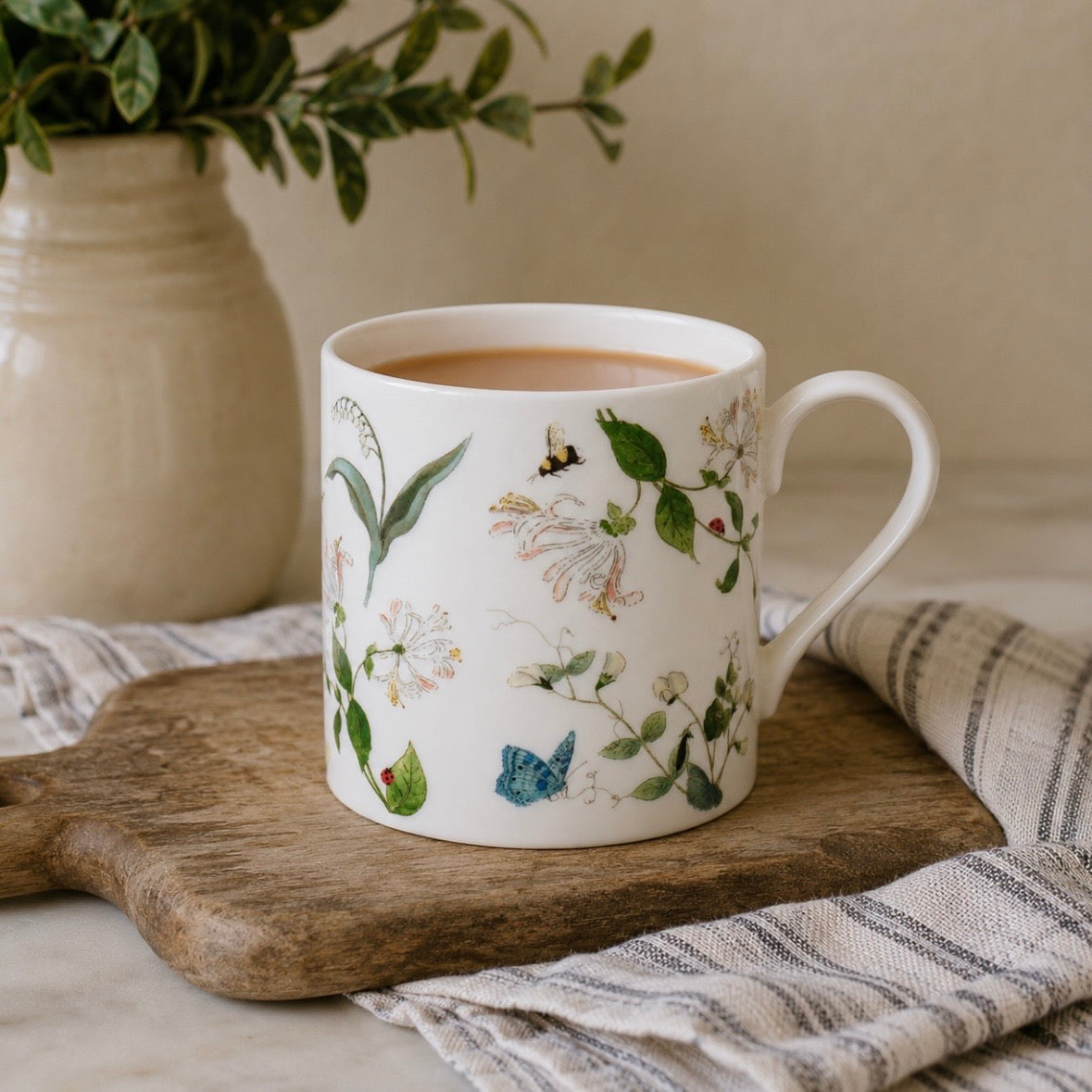 A white fine bone china mug decorated with delicate botanical illustrations of flowers, leaves, a bee, and a butterfly, styled on a wooden cutting board with a striped cloth and a potted plant in the background.