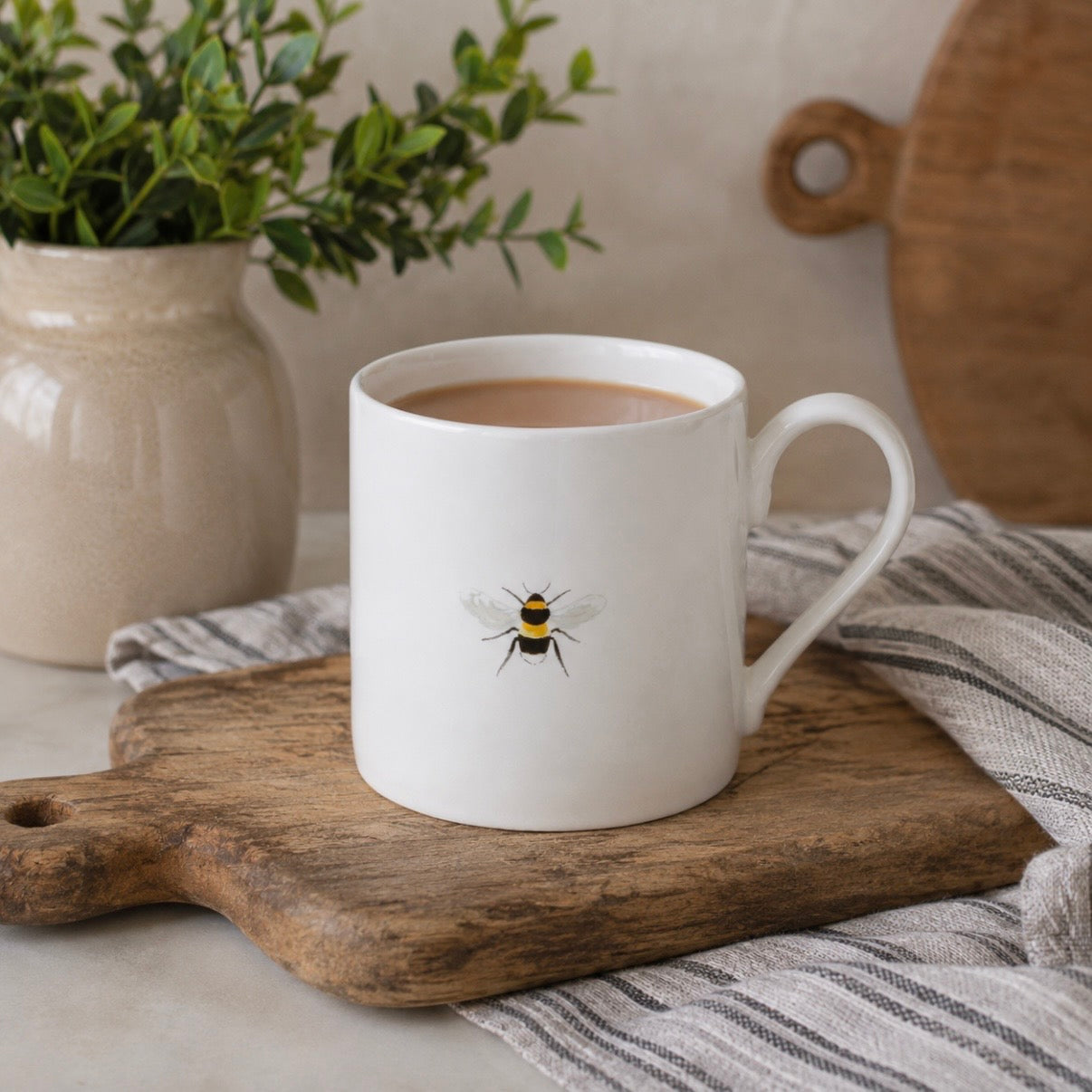 A white fine bone china mug with a handle, decorated with a single bee, resting on a wooden cutting board with a striped cloth underneath.