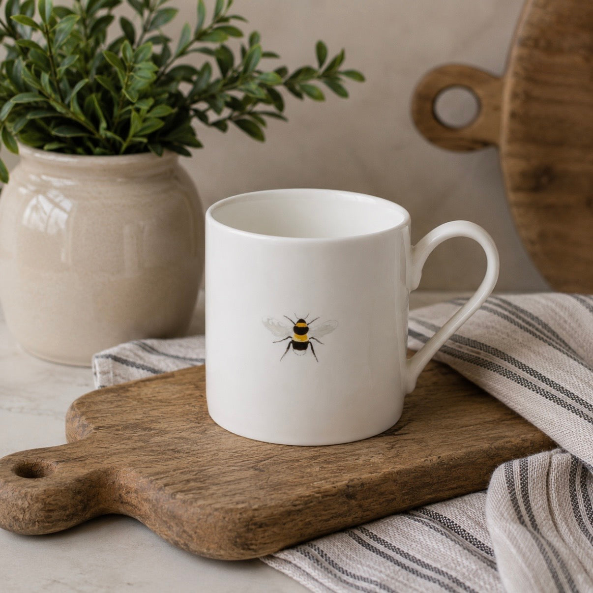 A white fine bone china mug with a handle, decorated with a single bee, resting on a wooden cutting board with a striped cloth underneath.