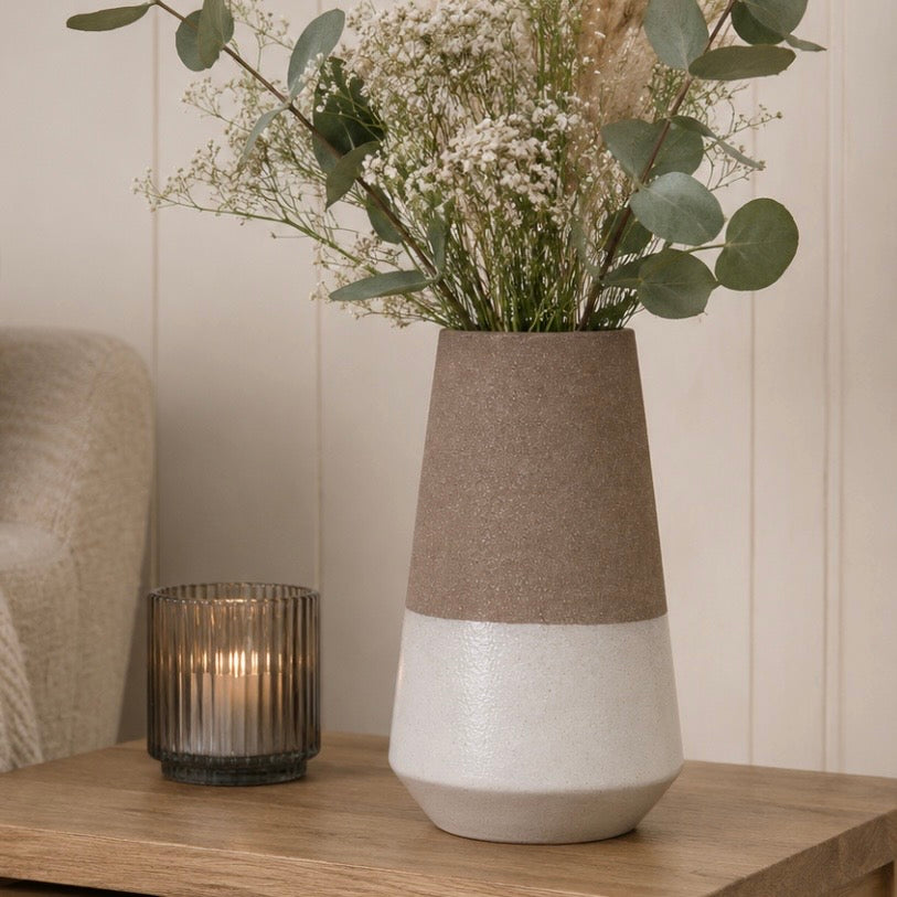 A two-tone ceramic vase with a textured mocha-brown top half and a gloss off-white bottom half, filled with dried flowers and greenery, sitting on a wooden table next to a lit candle in a ribbed glass holder.