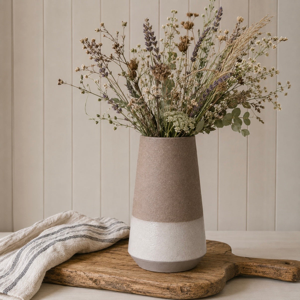 A two-tone ceramic vase with a textured matte finish, featuring a warm mocha brown upper half and a cream lower half, filled with dried flowers and placed on a rustic wooden board with a striped cloth against a beige paneled wall.