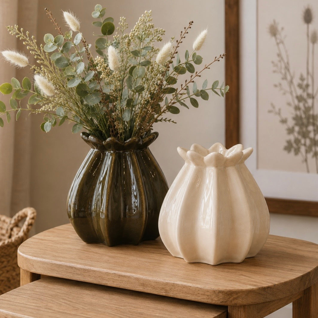 Two decorative ceramic poppy bud-shaped vases on a wooden table, one dark green and one cream, with dried grasses and eucalyptus stems in the larger vase, in a softly styled interior setting.