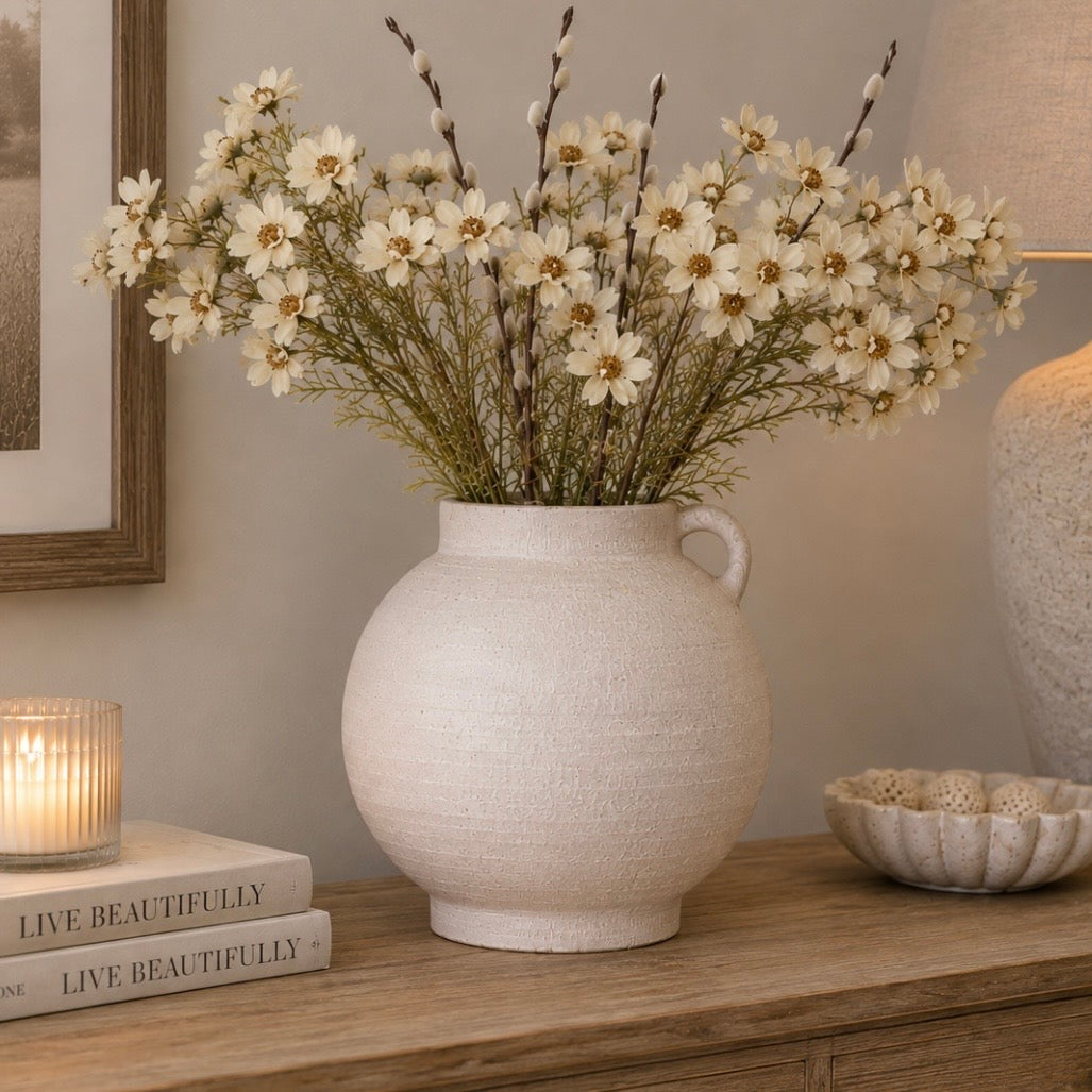 A textured cream terracotta vase filled with white daisy-like dried flowers and brown stems, displayed on a wooden console table in a styled room setting with a candle, books, and a lamp.