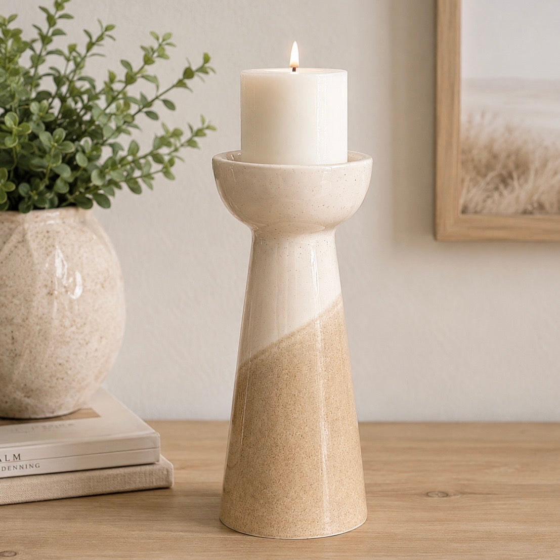 A ceramic candle holder with a tapered base, featuring a glossy off-white upper section and a textured beige lower section, holding a lit white pillar candle on a wooden tabletop.