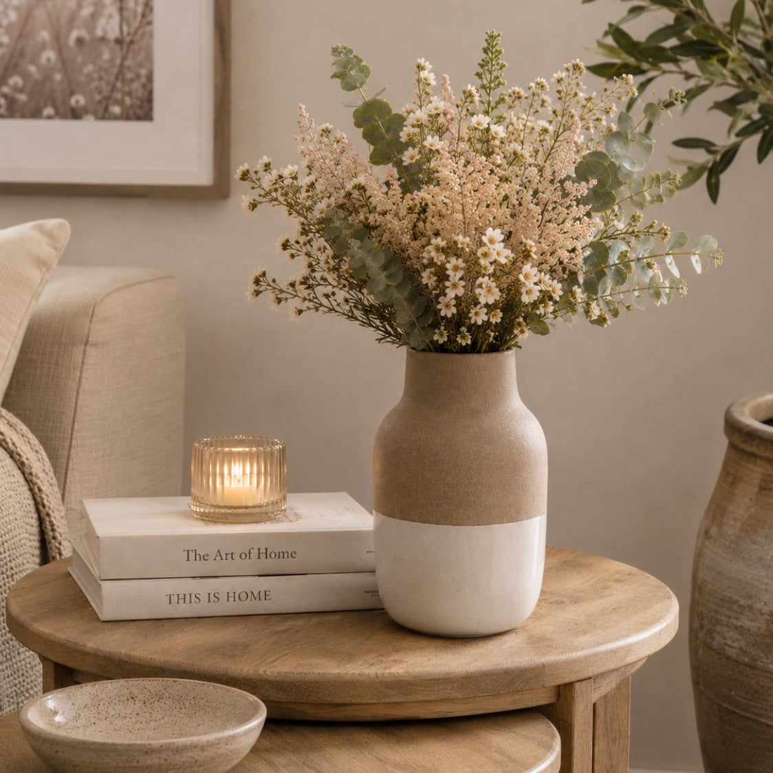 A two-tone ceramic vase with a matte beige top half and white bottom half, filled with dried white flowers and green eucalyptus stems, styled on a round wooden side table in a cozy living room setting.