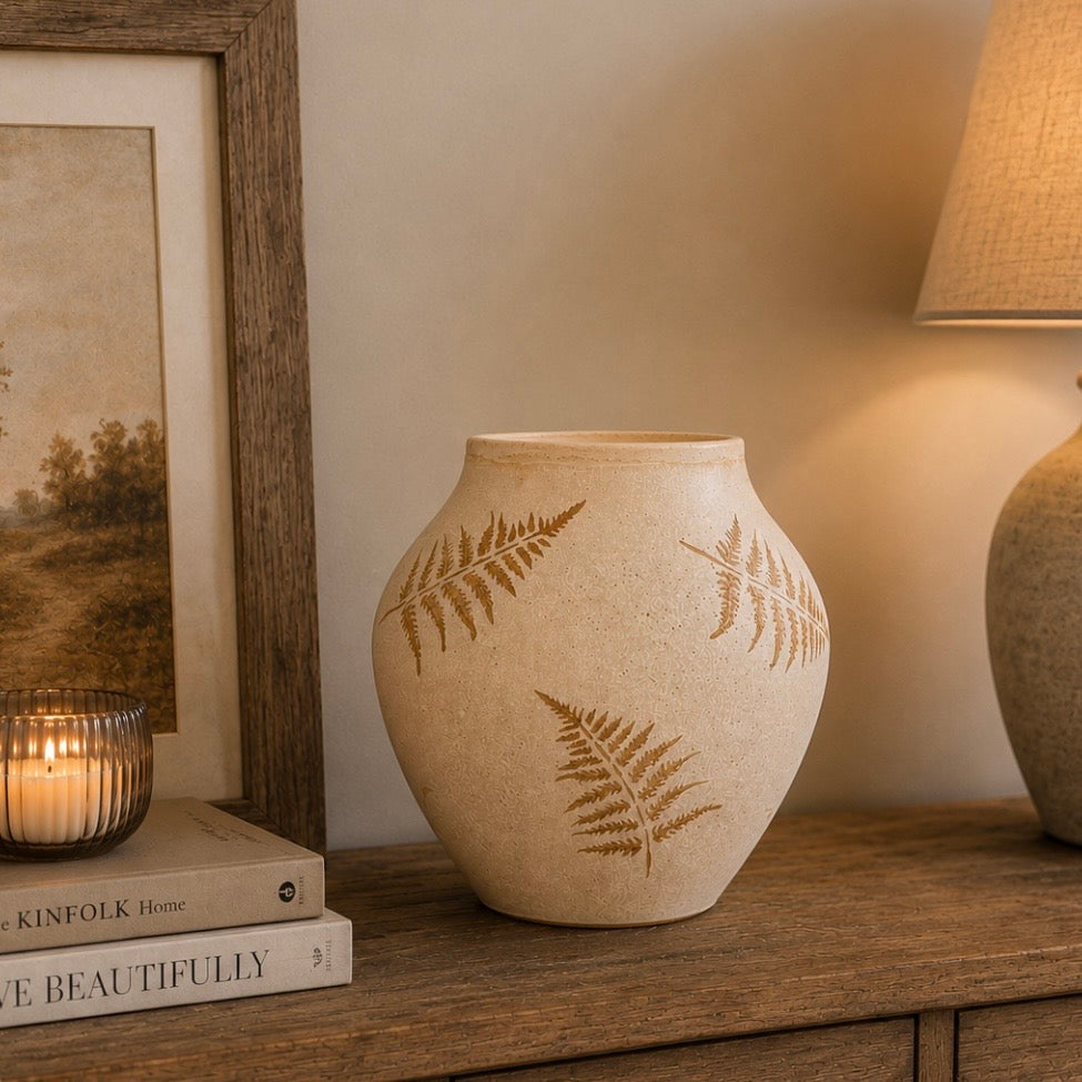 A beige ceramic vase with brown fern leaf patterns, styled on a wooden table next to a lit candle, framed picture, and a lamp.