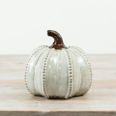 A decorative ceramic pumpkin with a glossy off-white glaze, vertical ribbed texture, beaded trim, and a dark brown stem, displayed on a light wooden surface against a white background.