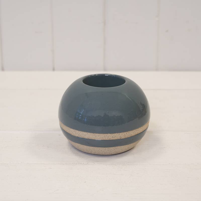 A small round ceramic vase with a glossy dark blue-gray finish and a textured beige band around the middle, set on a white wooden surface against a white paneled background.