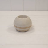 A small round ceramic vase with a glossy off-white finish and a textured beige band around the middle, set on a white wooden surface against a white paneled background.