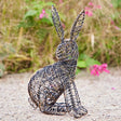 A wire-wrapped hare figurine in a sitting position, set outdoors on a gravel path with a blurred natural background of green foliage and pink flowers.