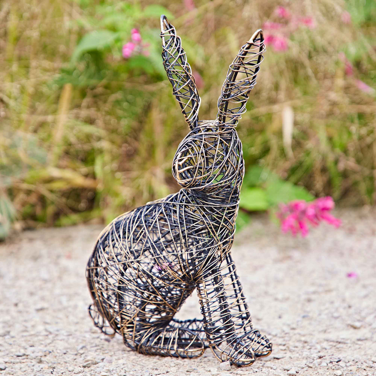 A wire-wrapped hare figurine in a sitting position, set outdoors on a gravel path with a blurred natural background of green foliage and pink flowers.