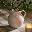White ceramic pitcher with a plant on a checkered tablecloth with a candle in the foreground.