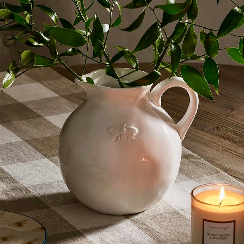 White ceramic pitcher with a plant on a checkered tablecloth with a candle in the foreground.