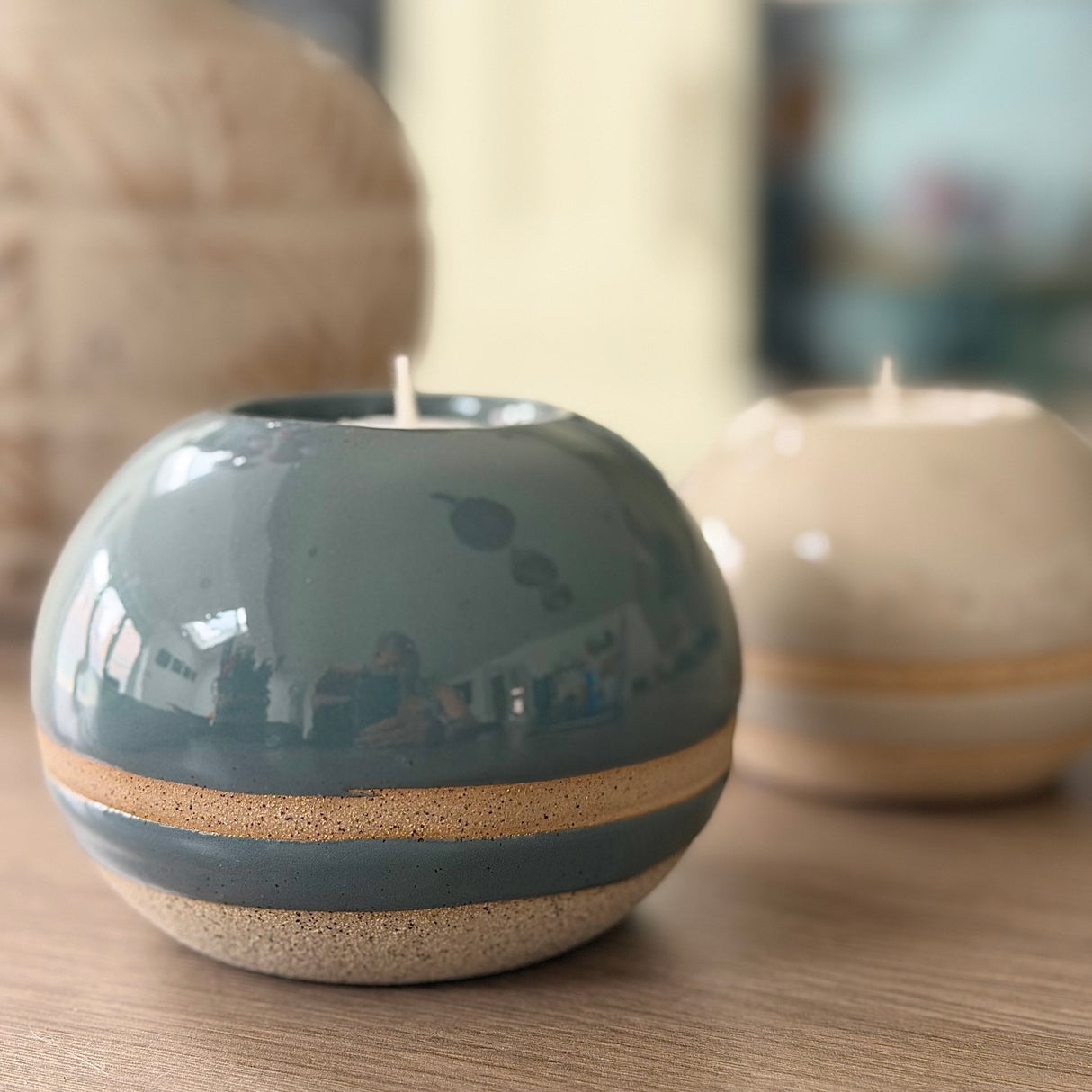 A round ceramic candle holder with a glossy slate blue top and a speckled beige base, holding a small white candle, set on a wooden surface with another blurred candle in the background.