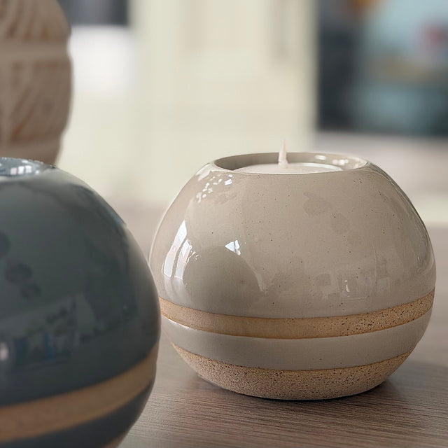 A small round ceramic candle holder with a white candle inside, featuring a glossy cream upper section, a textured beige band, and a speckled beige base, set on a wooden surface.