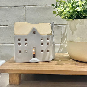 Ceramic house-shaped lantern with a candle inside on a wooden surface, next to a potted plant.