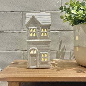 Ceramic house-shaped lantern with glowing interior on a wooden surface against a stone wall background.