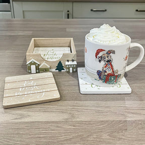 Mug with hot chocolate and whipped cream on a coaster, next to a decorative wooden box and coasters with Christmas-themed designs.