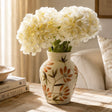 A cream ceramic vase with a orange and green floral pattern, filled with large white hydrangea blooms, placed on a wooden table in a softly lit room.
