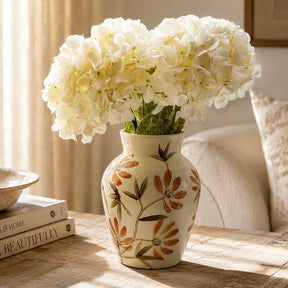 A cream ceramic vase with a orange and green floral pattern, filled with large white hydrangea blooms, placed on a wooden table in a softly lit room.