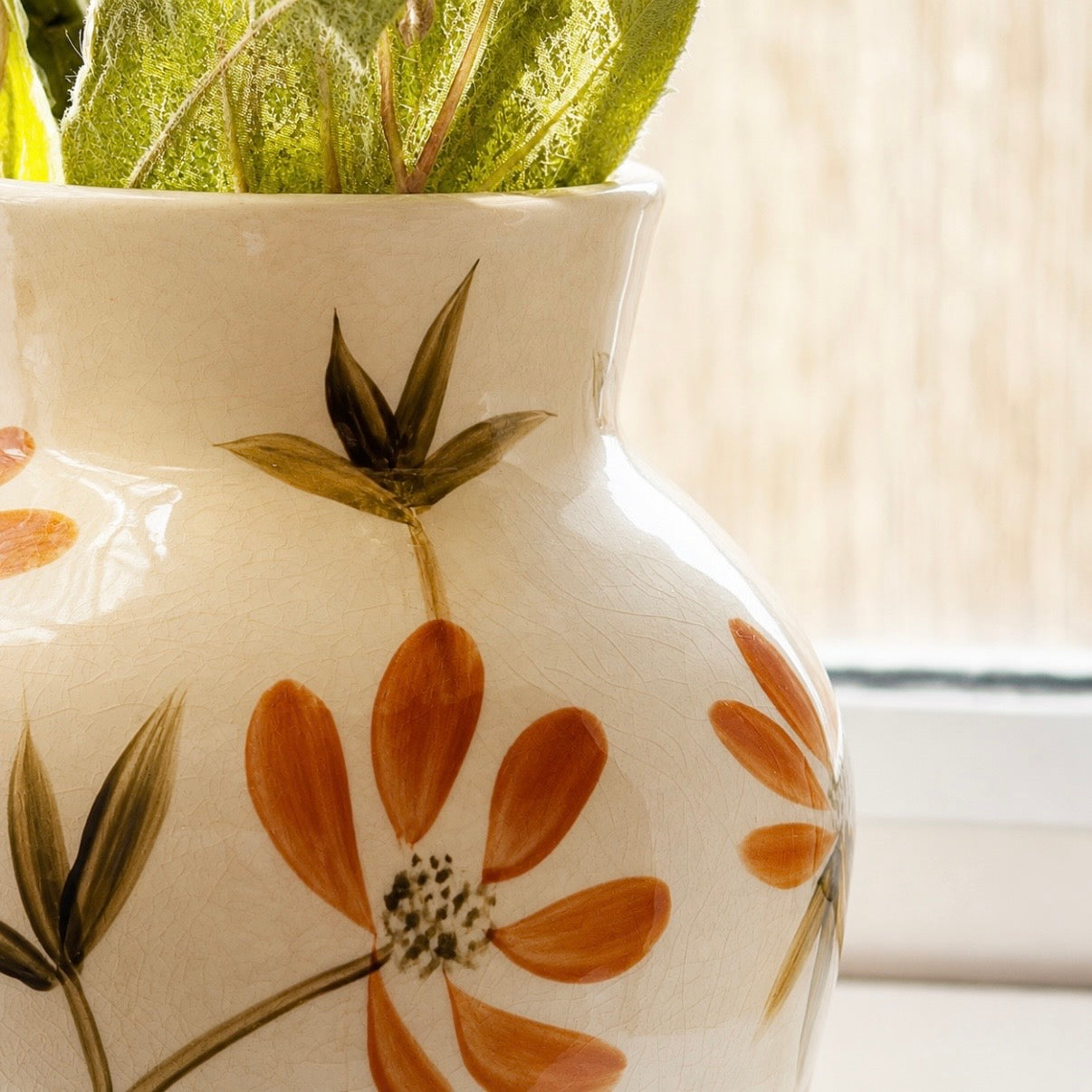 A close-up of a glossy cream ceramic vase with hand-painted orange flowers and green leaves, holding green foliage, set against a softly blurred window background.