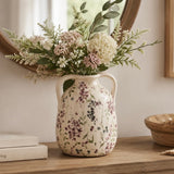 A cream-colored ceramic vase with a floral pattern and two side handles, filled with a bouquet of white and pink faux flowers and green foliage, displayed on a wooden table in a styled interior setting.