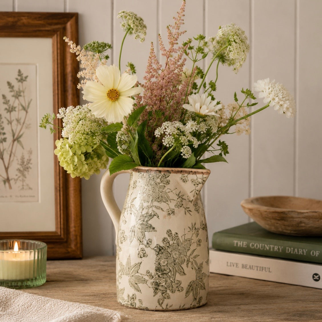 A ceramic pitcher vase with a green floral toile pattern on a cream background, filled with a bouquet of white and pale green wildflowers, set on a wooden table next to a candle and books.