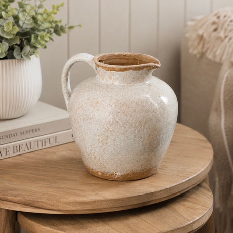 A ceramic grey pitcher vase with a rounded body, small handle, and wide flared rim, shown on a wooden table in a styled interior setting with books, a plant, and a woven chair visible.