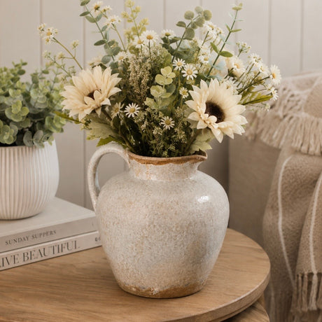 A rustic grey ceramic pitcher vase filled with a bouquet of artificial flowers, including sunflowers and daisies, styled on a wooden table next to a potted plant and a cozy throw blanket.