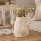A rustic ceramic pitcher vase filled with dried white flowers, placed on a wooden table in a softly lit living room setting.
