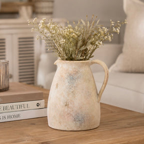 A rustic ceramic pitcher vase filled with dried white flowers, placed on a wooden table in a softly lit living room setting.