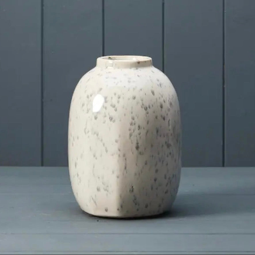 A ceramic vase with a rounded, bulbous shape and a glossy off-white glaze speckled with grey, set against a dark grey paneled background.