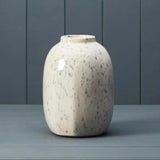 A ceramic vase with a rounded, bulbous shape and a glossy off-white glaze speckled with grey, set against a dark grey paneled background.