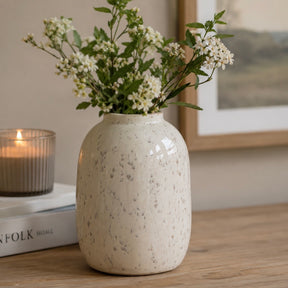 A speckled white and grey ceramic vase filled with small white flowers and green stems, placed on a wooden table beside a lit candle and a framed picture in a softly lit room.