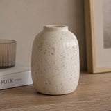 A small ceramic vase with a textured, speckled white and grey finish, standing on a wooden tabletop next to a book and a glass candle holder.