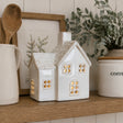 A small white ceramic house-shaped candle holder with glowing warm lights inside, displayed on a wooden shelf alongside a mug, spoon, framed botanical print, and a potted plant.
