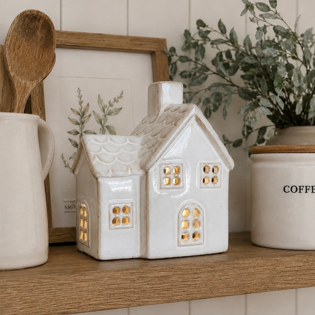 A small white ceramic house-shaped candle holder with glowing warm lights inside, displayed on a wooden shelf alongside a mug, spoon, framed botanical print, and a potted plant.