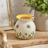 A ceramic wax burner with a sunflower-shaped top, decorated with bumblebees and small wildflowers on a white background, styled on a wooden surface with a potted plant and framed picture in the background.