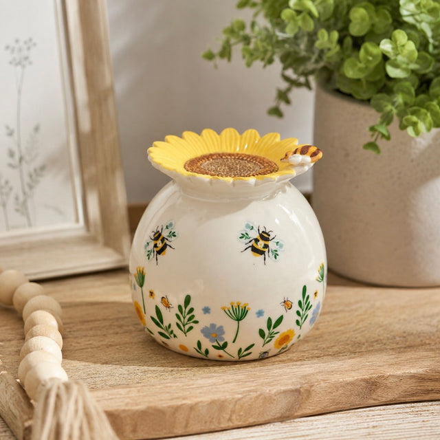 A ceramic wax burner with a sunflower-shaped top, decorated with bumblebees and small wildflowers on a white background, styled on a wooden surface with a potted plant and framed picture in the background.