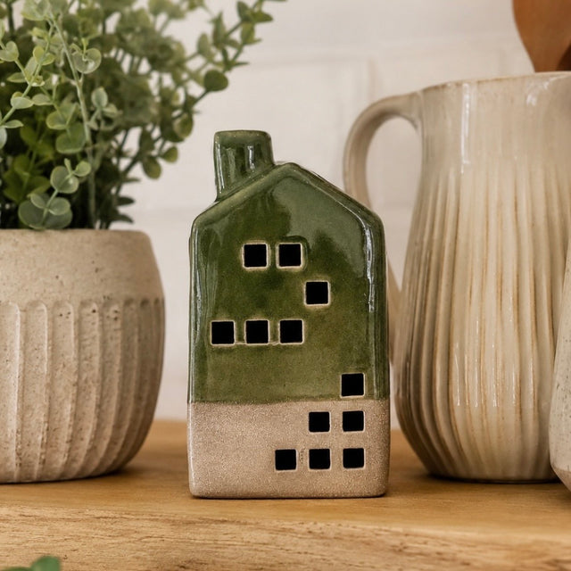 A small glossy ceramic house-shaped candle holder or decor piece in olive green and beige, shown on a wooden surface with a potted plant and a ribbed ceramic pitcher in the background.