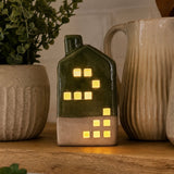 A small ceramic house-shaped LED tea light with a dark green upper half and beige lower half, glowing with warm yellow window lights, displayed on a wooden surface between ceramic mugs and a potted plant.