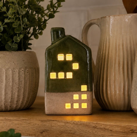 A small ceramic house-shaped LED tea light with a dark green upper half and beige lower half, glowing with warm yellow window lights, displayed on a wooden surface between ceramic mugs and a potted plant.
