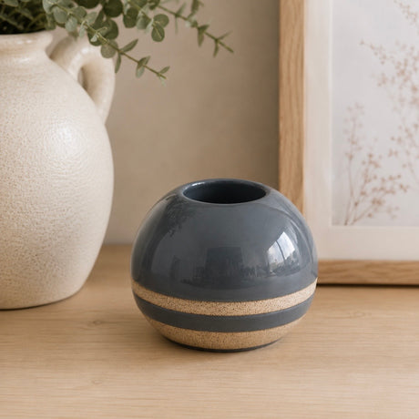 A small round ceramic tealight holder in blue with a horizontal beige band near the base, shown on a wooden surface with a larger cream vase and framed botanical art in the background.