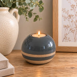 A round ceramic tea light holder with a blue top and a textured beige lower band, holding a lit tealight candle on a wooden table with home decor props.