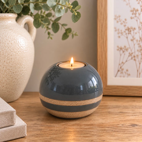 A round ceramic tea light holder with a blue top and a textured beige lower band, holding a lit tealight candle on a wooden table with home decor props.