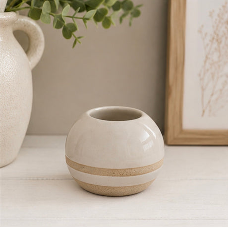 A small round ceramic tealight holder with a glossy off-white finish and a textured beige band around the middle, displayed on a white tabletop with a neutral background and home decor props.